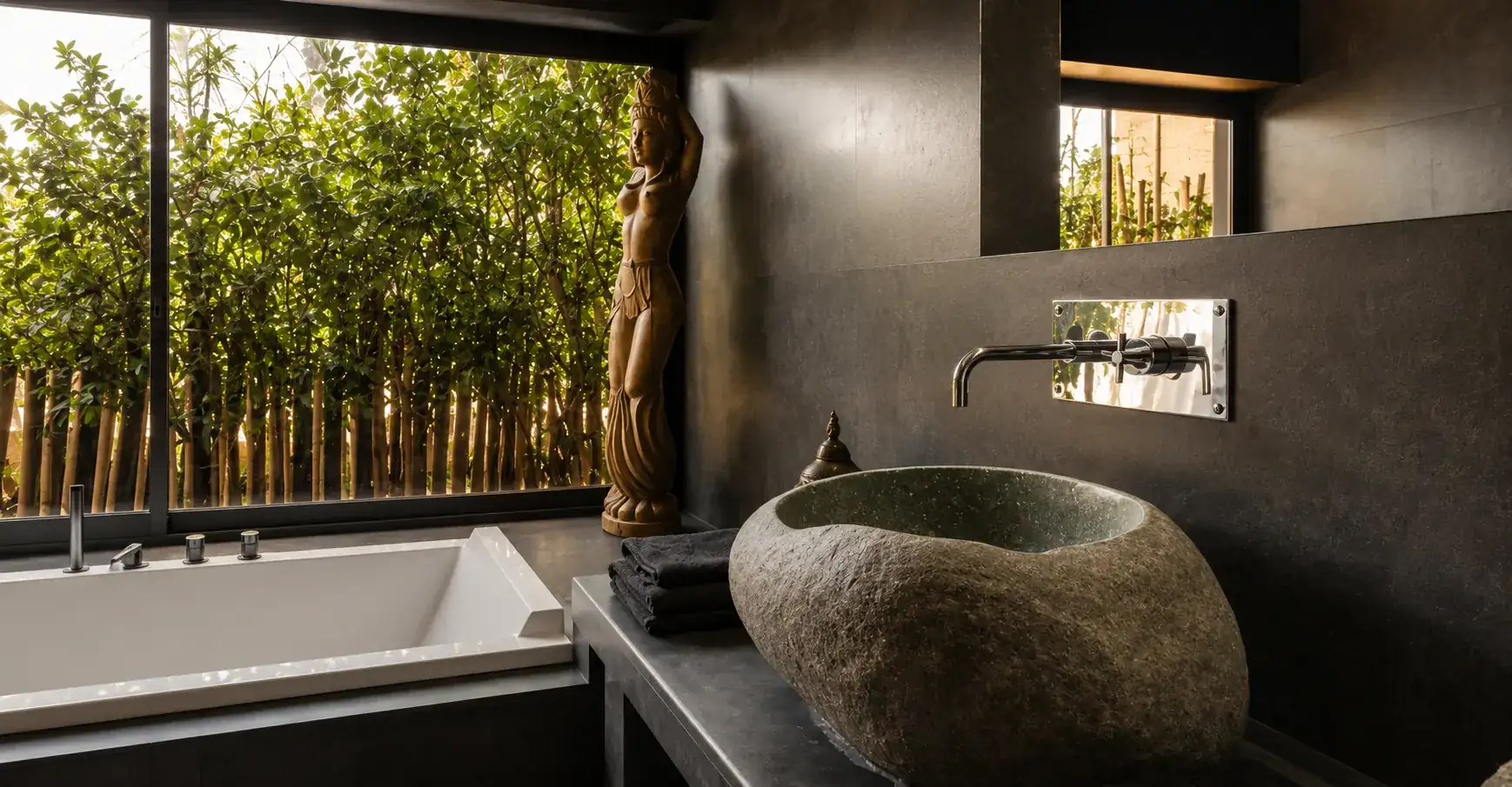 Vasque en galet brut et statue de bois dans une salle de bain de la Villa Nausikaa, ouverte sur la verdure