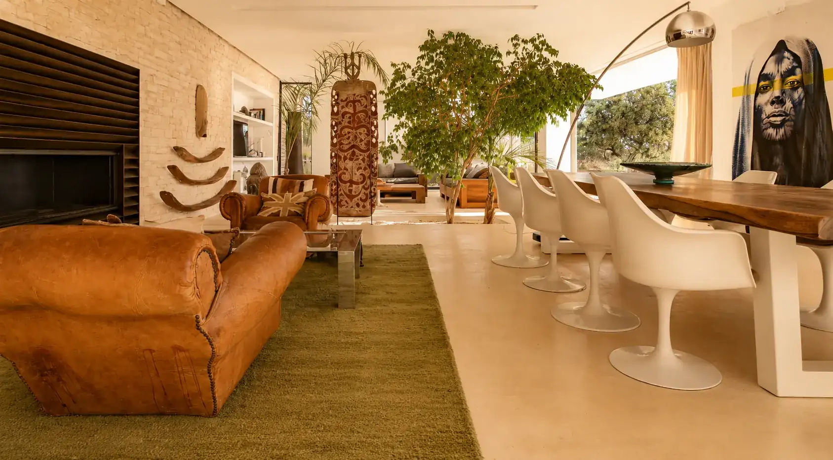 Cheminée murale, canapé en cuir patiné et table d'hôtes entourée de chaises Tulip blanches à la Villa Nausikaa